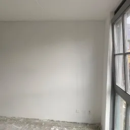 Świeżo wykończone, białe ściany w pustym pokoju z dużym oknem i widocznymi gniazdkami elektrycznymi; podłoga z resztkami zaprawy.