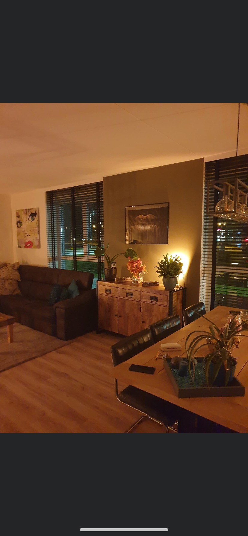 Umeblowane wnętrze salonu z brązową sofą, drewnianą komodą z roślinami, drewnianym stołem z dekoracjami i obrazami na ścianach w ciepłym oświetleniu.