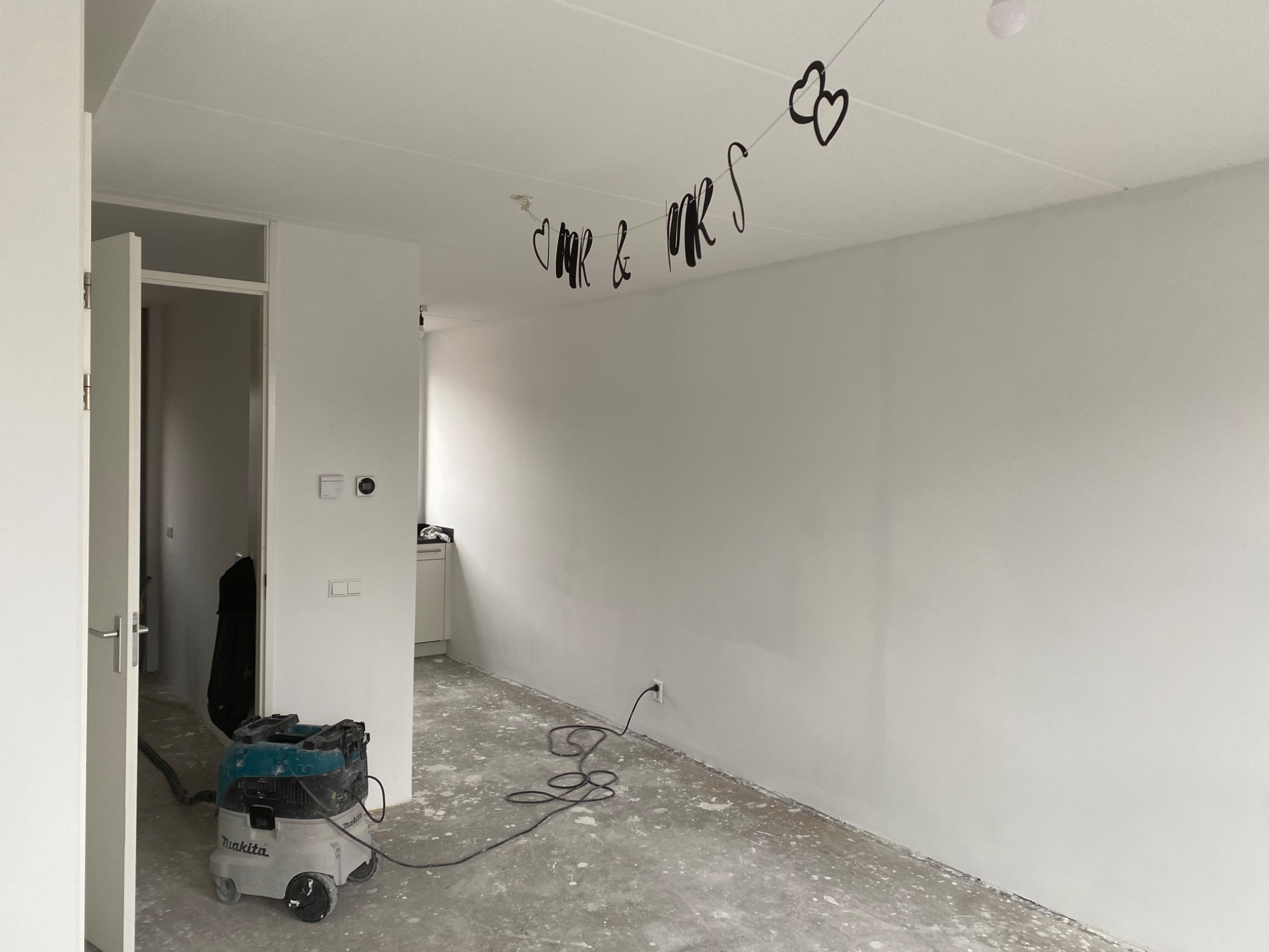 Wnętrze po świeżym malowaniu na biało, z dekoracją 'Mr & Mrs' pod sufitem i odkurzaczem przemysłowym Makita w rogu pokoju.