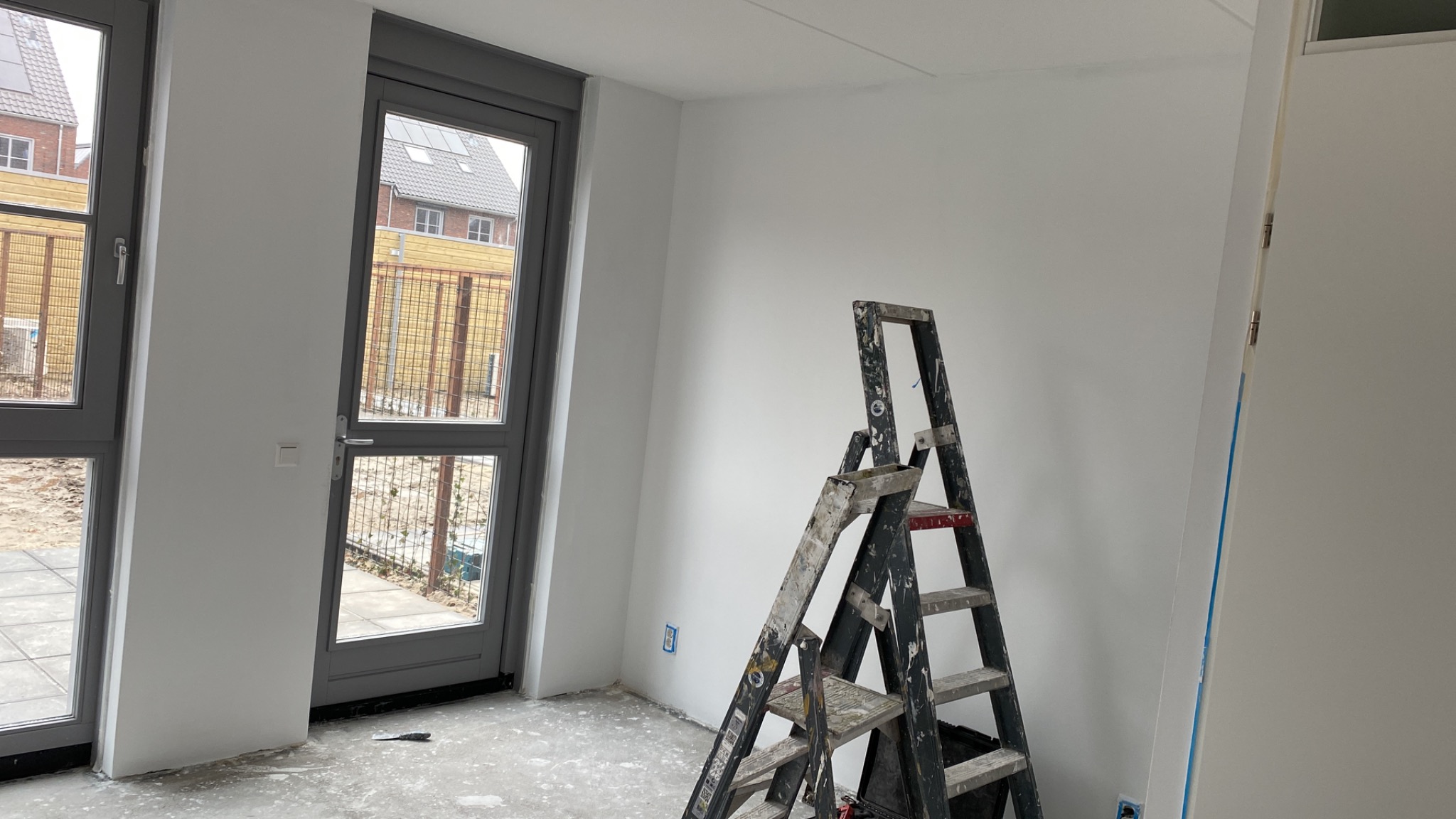 Pokój w trakcie prac wykończeniowych: świeżo pomalowane białe ściany, aluminiowa drabina oparta o ścianę, widoczne ślady farby na podłodze, szare ramy okienne i drzwiowe.