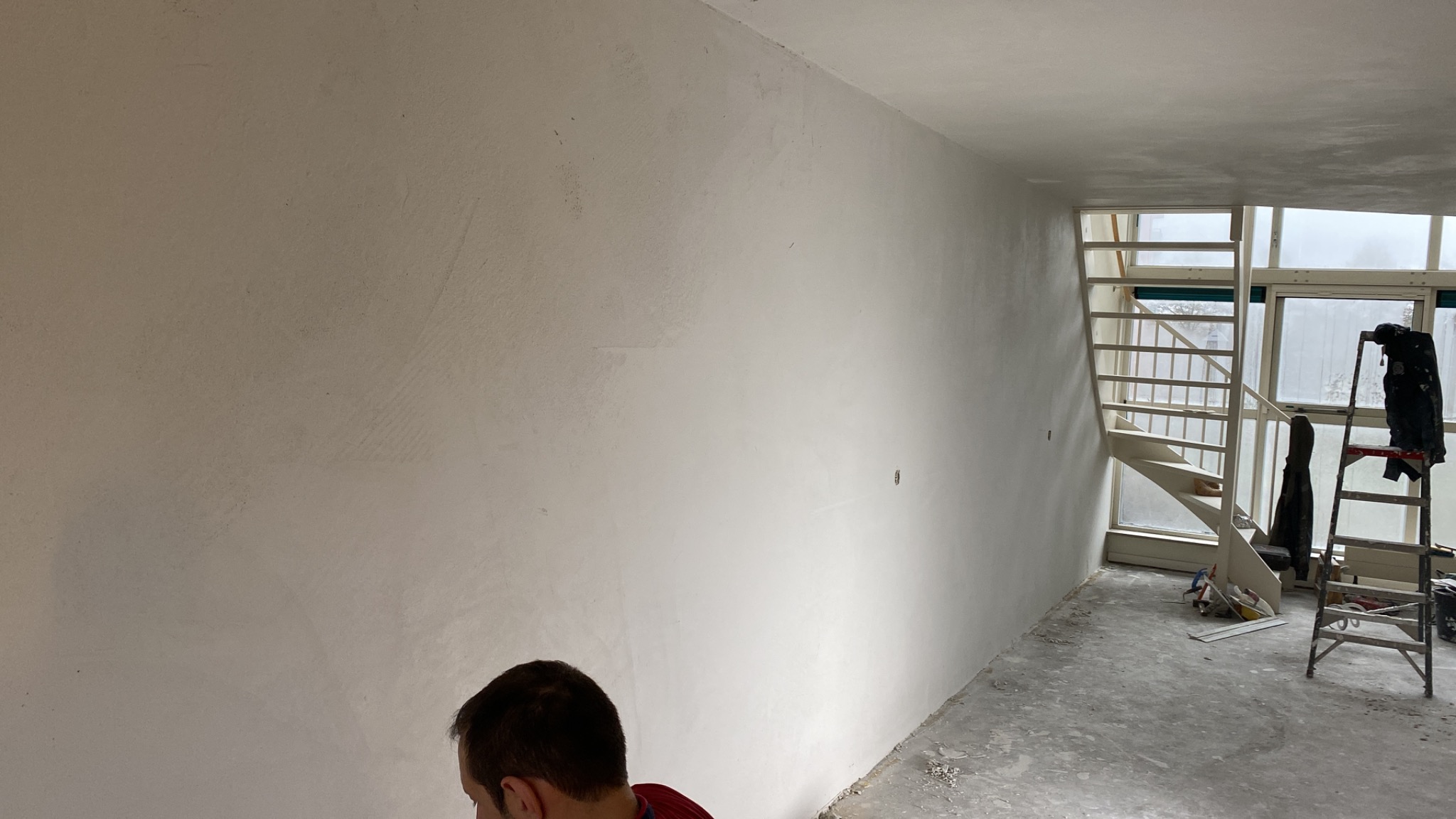 Świeżo otynkowana ściana w jasnym pomieszczeniu, widoczne ślady po szpachlowaniu, drabina oparta o okno, przygotowania do dalszych prac wykończeniowych.