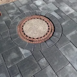 Szare kostki ułożone wokół okrągłej studzienki kanalizacyjnej z żeliwną pokrywą, widok z góry.