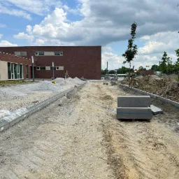 Przygotowanie terenu pod nawierzchnię: wyrównywanie podłoża, ustawianie obrzeży, hałdy żwiru, maszyna do ubijania ziemi i paleta z kostką na placu budowy przy budynku.