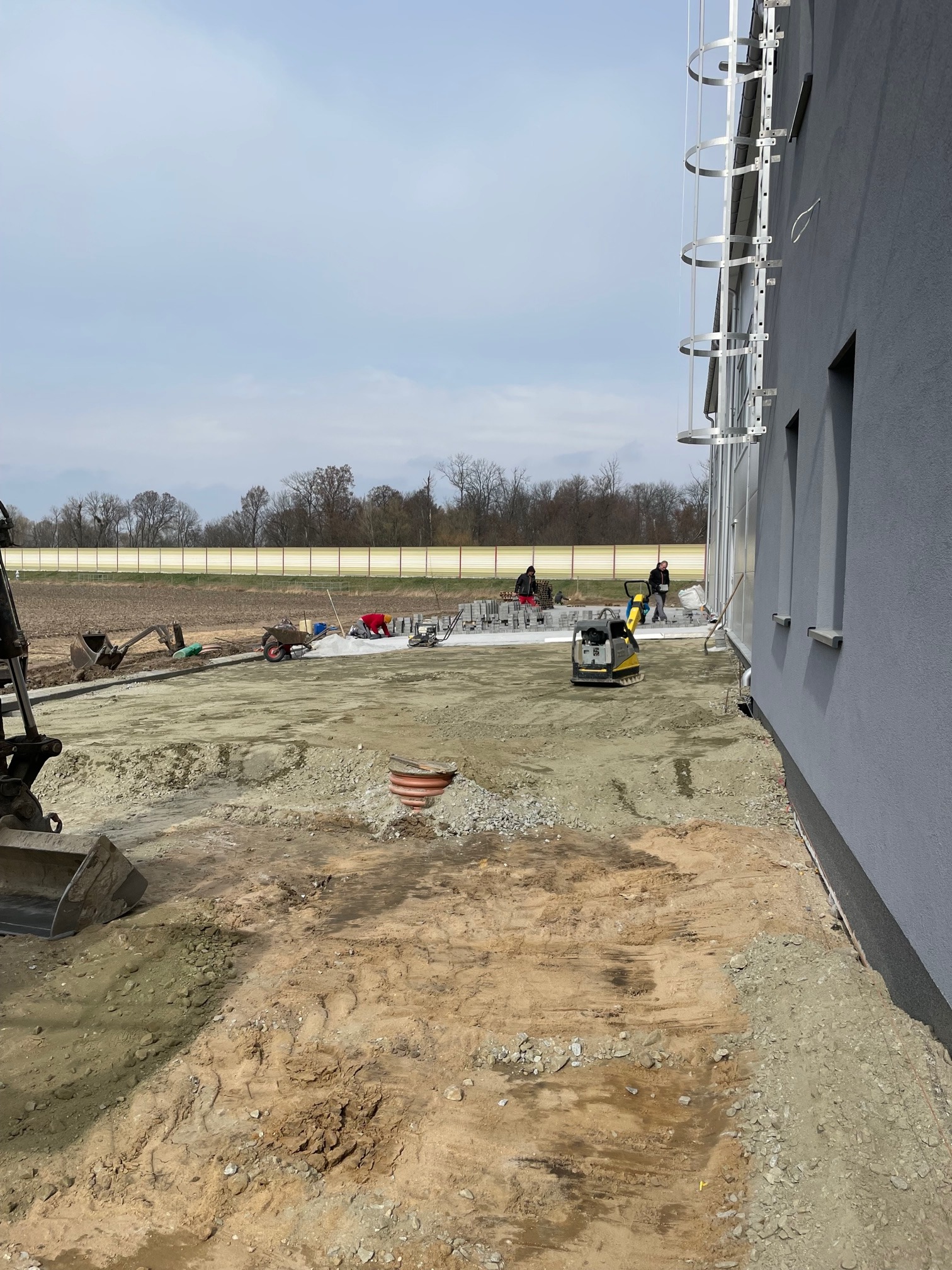 Przygotowanie terenu pod budowę, widoczni robotnicy, zagęszczarka, koparka i inne maszyny budowlane. Na gruncie widoczne ślady opon. Prace ziemne w toku.