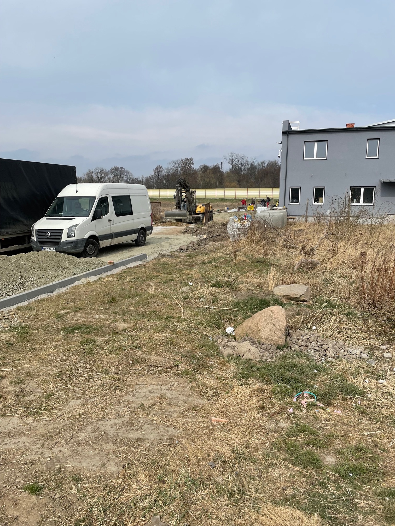 Układanie obrzeży betonowych przy nowo powstającym parkingu, w tle ekipa budowlana i mini koparka.