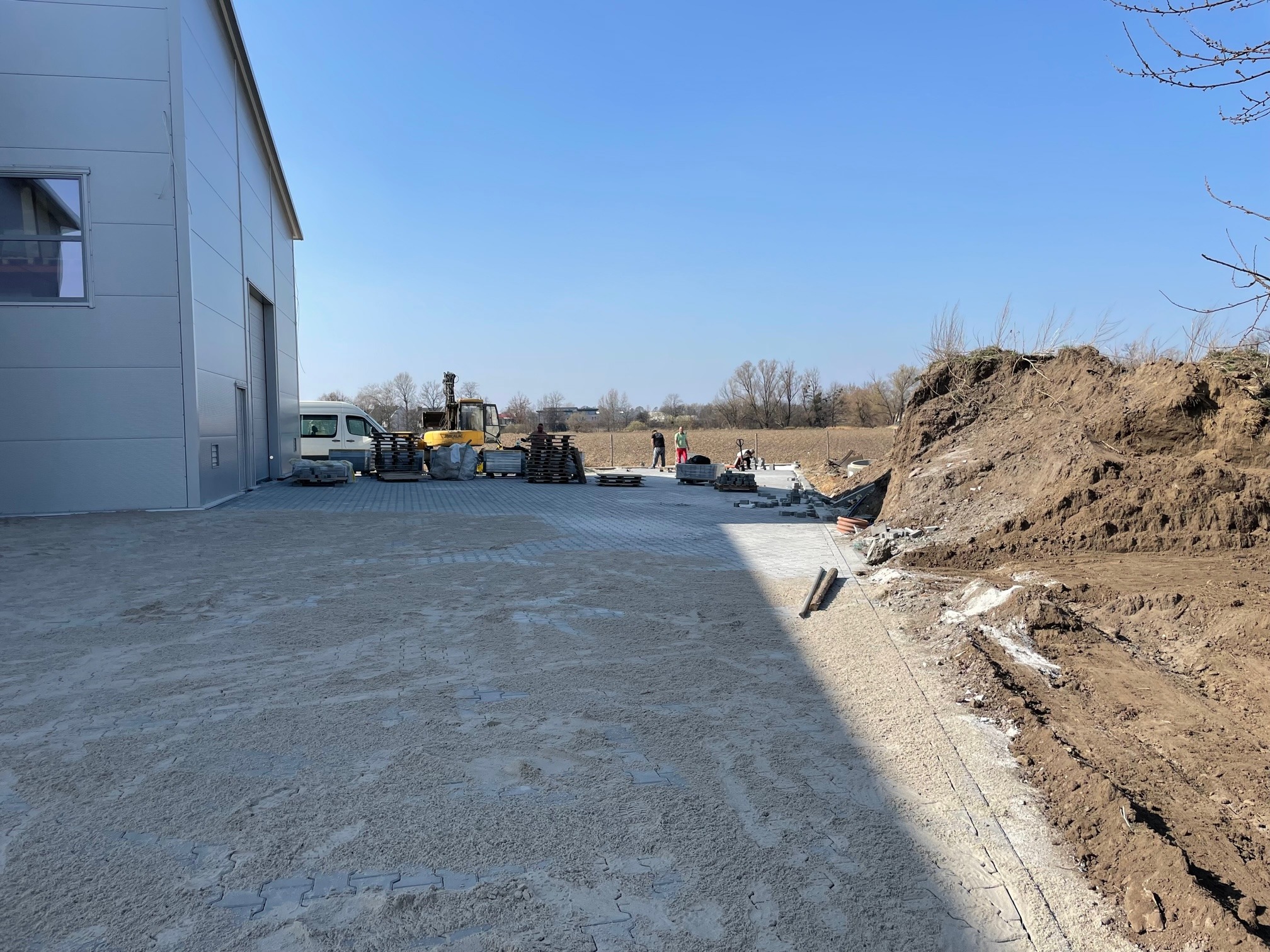 Plac budowy z częściowo ułożoną kostką brukową, widoczny budynek magazynowy, koparka oraz pracownicy w oddali.