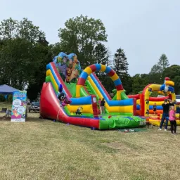Kolorowy dmuchany zamek ze zjeżdżalniami i motywami zwierząt, ustawiony na trawniku podczas imprezy plenerowej, w tle widoczny namiot i dzieci.