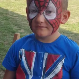 Chłopiec z pomalowaną twarzą w motywie Spider-Mana, siedzący na krześle na tle zielonej trawy i rozmytych postaci.