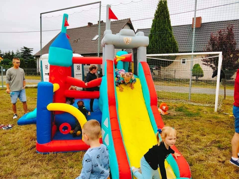 Dzieci bawią się na dmuchanym zamku ze zjeżdżalnią w kształcie twierdzy, ustawionym na trawniku przed domem i ogrodzeniem z siatki.