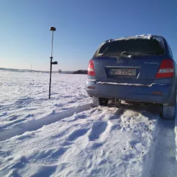 Niebieska Kia Sorento z numerem rejestracyjnym NP1 22542 zaparkowana na zasypanym śniegiem polu, obok stoi geodezyjny odbiornik GPS na tyczce.