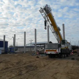 Żuraw samochodowy podczas montażu prefabrykowanych słupów na placu budowy, widoczne ślady opon na piaszczystym podłożu i błękitne niebo z chmurami w tle.