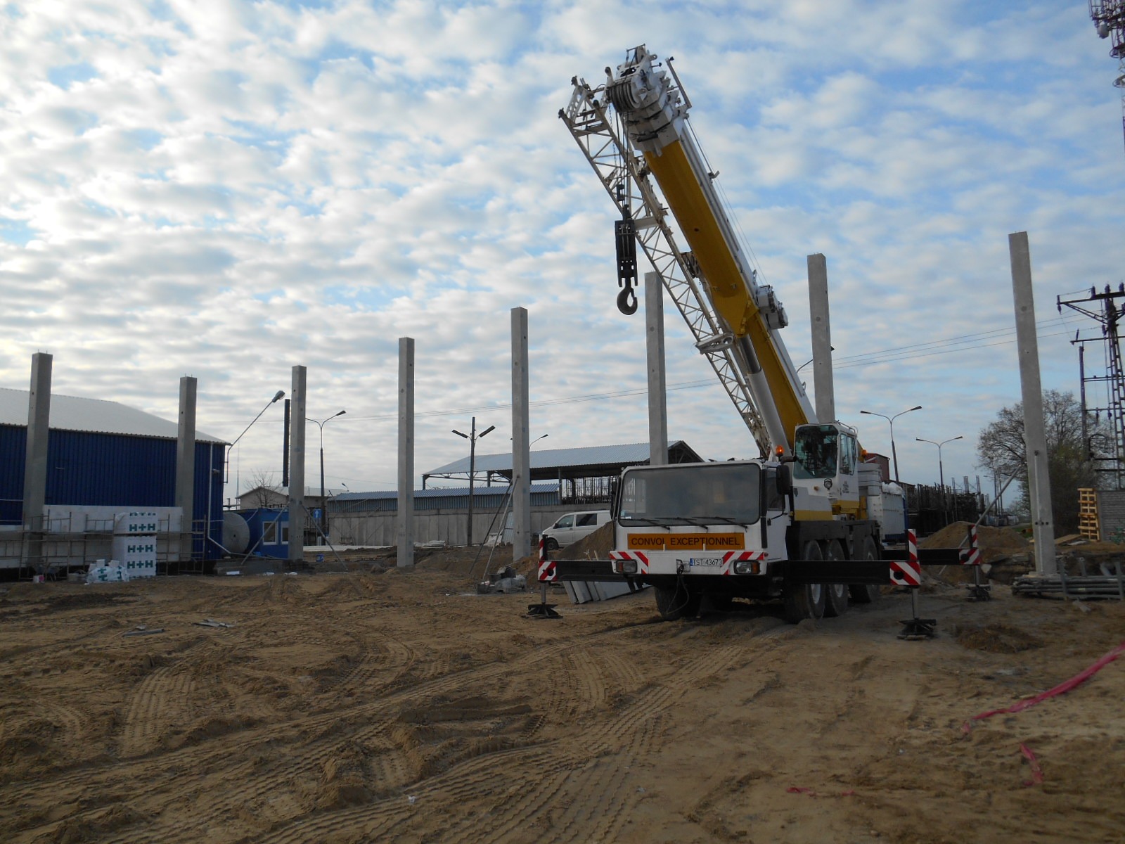 Żuraw samochodowy podczas montażu prefabrykowanych słupów na placu budowy, widoczne ślady opon na piaszczystym podłożu i błękitne niebo z chmurami w tle.