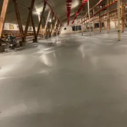 Świeżo wylana, gładka, szara posadzka w hali przemysłowej, widoczna maszyna do zacierania betonu, drewniane słupy podtrzymujące konstrukcję dachu i czerwone karmniki dla zwierząt.