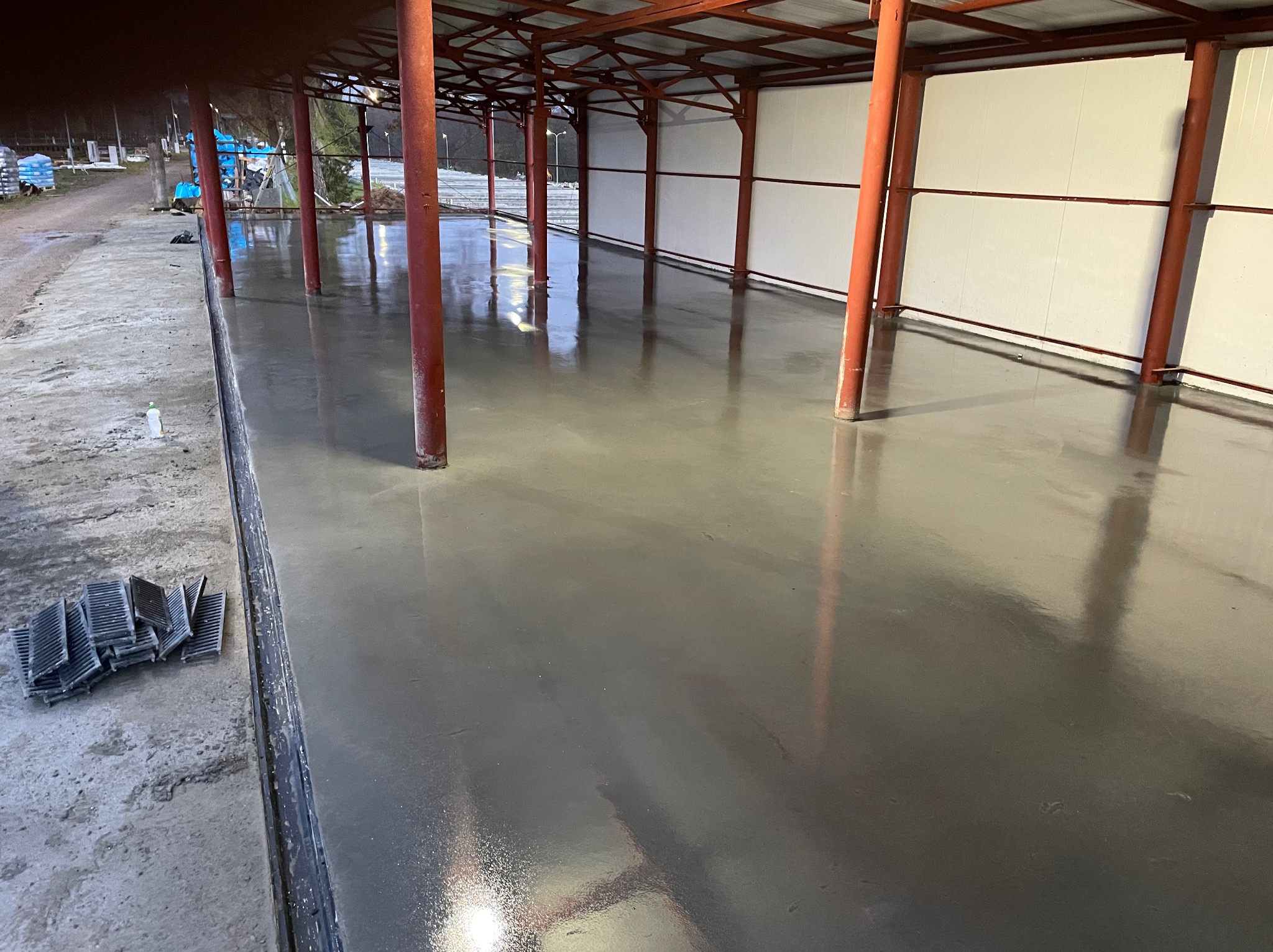 Świeżo wylana, gładka, szara posadzka w hali z czerwonymi słupami nośnymi i białymi ścianami. Na krawędzi posadzki leżą czarne elementy kratki ściekowej.