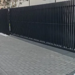 Nowoczesna, antracytowa brama wjazdowa z pionowych profili, uzupełniona ogrodzeniem o tej samej stylistyce. Białe kamienie dekoracyjne wzdłuż podstawy. Kostka brukowa na podjeździe.