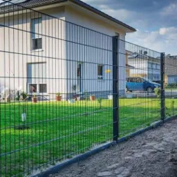 Nowoczesne ogrodzenie panelowe w kolorze grafitowym, widoczne na tle domu z zadbanym trawnikiem i błękitnym samochodem w oddali, z delikatnym pochyleniem kamery w prawo.