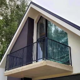 Nowoczesny balkon z czarną metalową balustradą przy domu o trójkątnym dachu, okno zabezpieczone folią ochronną.