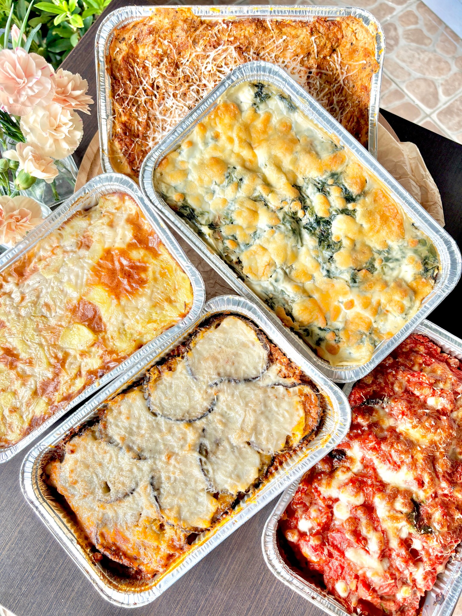 Pięć aluminiowych pojemników z różnymi rodzajami zapiekanek: mięsna posypana serem, szpinakowa pod beszamelem, warzywna z ziemniakami, bakłażanowa z mozzarellą i pomidorowa z bazylią, na drewnianym...