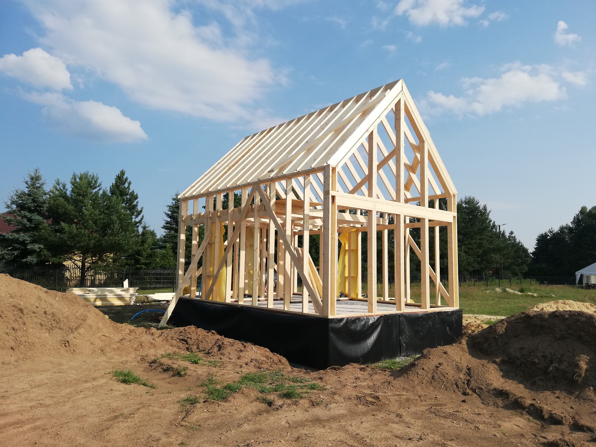 Drewniany szkielet powstającego domu jednorodzinnego na fundamencie z izolacją, widok z zewnątrz na tle błękitnego nieba i zieleni drzew.