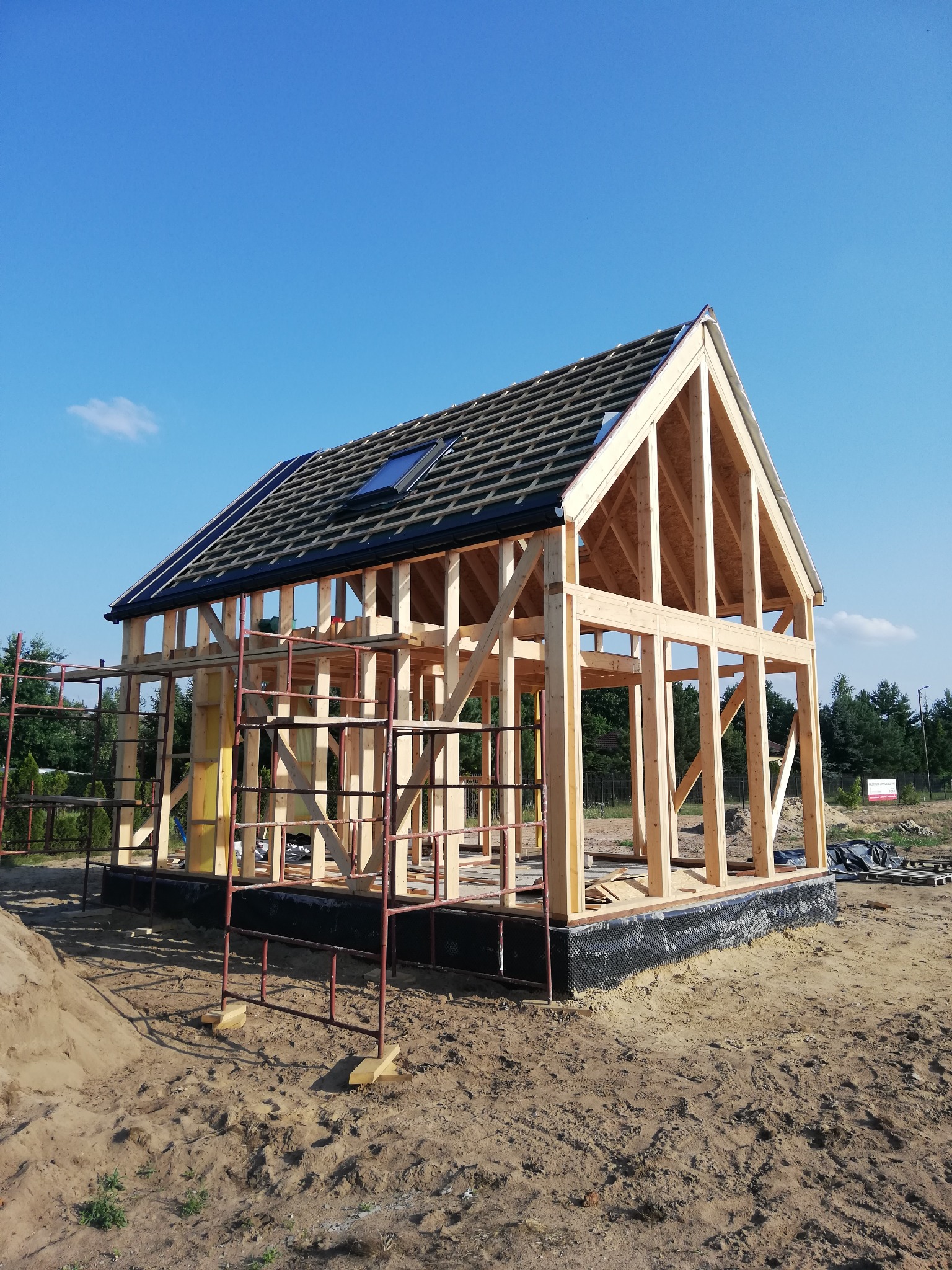Szkielet drewniany domu w trakcie budowy, widoczny fundament z izolacją, rusztowanie oraz dach z oknem połaciowym na tle błękitnego nieba.