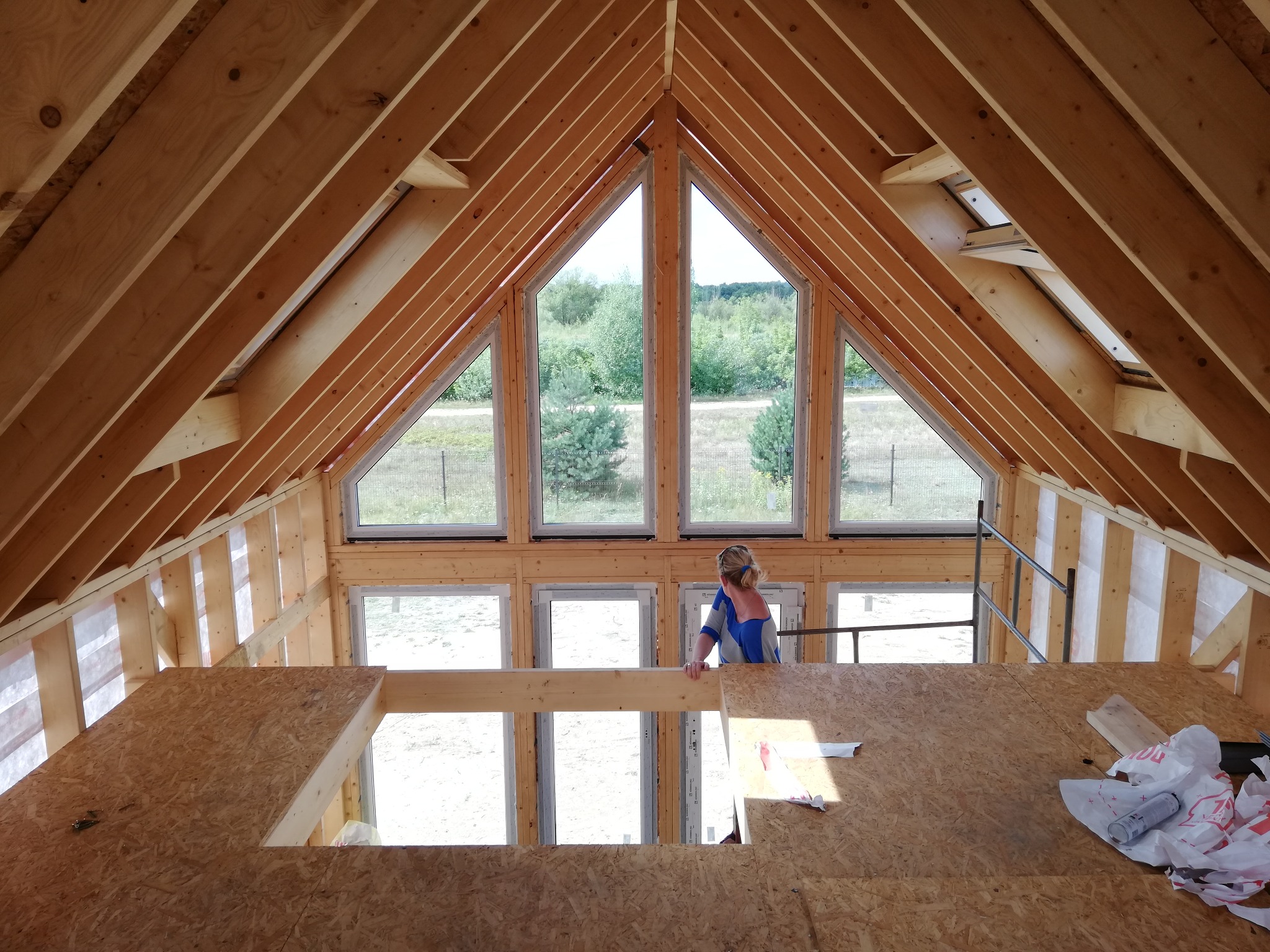 Wnętrze domu w trakcie budowy z widoczną konstrukcją drewnianą dachu, okna szczytowe z widokiem na zieleń, oraz kobieta w niebieskiej koszulce stojąca na podwyższeniu z płyty OSB.
