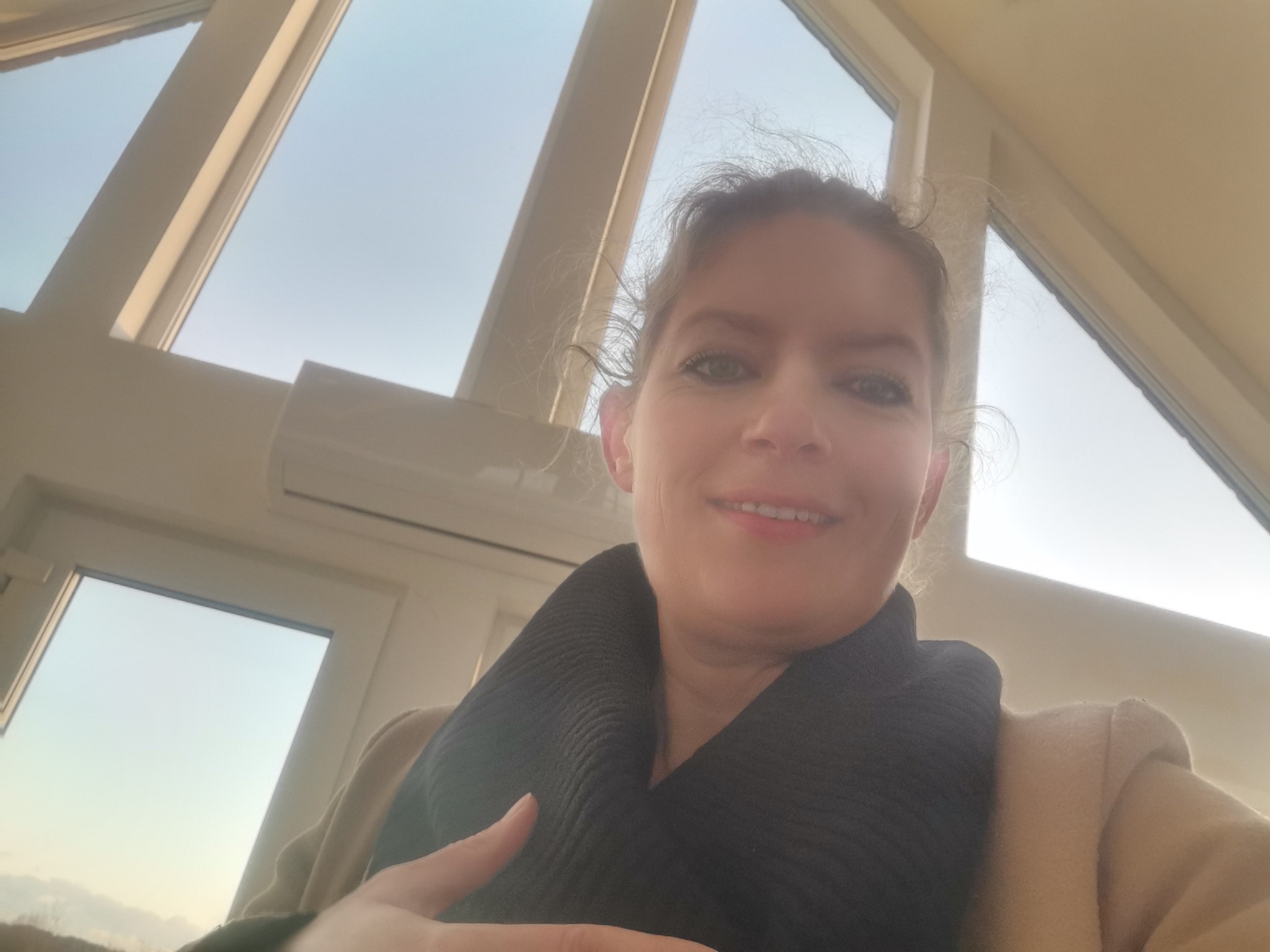 Portret kobiety w beżowym płaszczu i czarnym szaliku na tle okna dachowego z widokiem na jasne niebo.