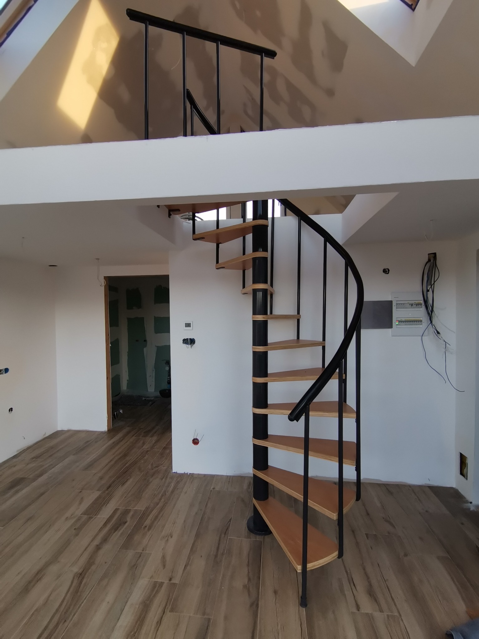 Wnętrze z drewnopodobną podłogą i spiralnymi schodami z czarną metalową konstrukcją i drewnianymi stopniami, widok na antresolę i niedokończone pomieszczenie w tle.