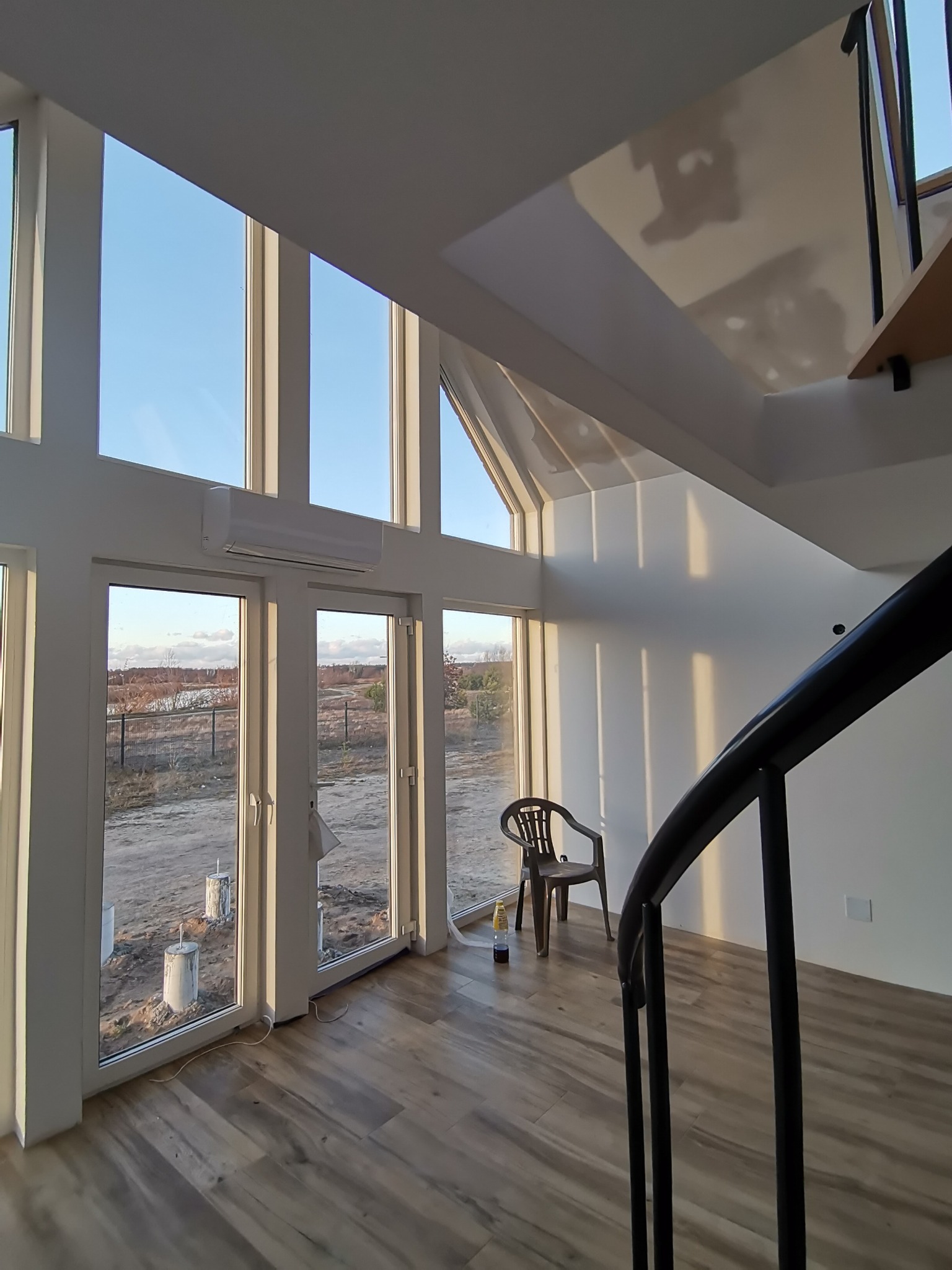 Nowoczesne wnętrze z dużymi oknami, przez które wpada popołudniowe światło, oświetlając drewnianą podłogę i białe ściany. Widoczna czarna, spiralna balustrada schodów i krzesło z butelką płynu obok.