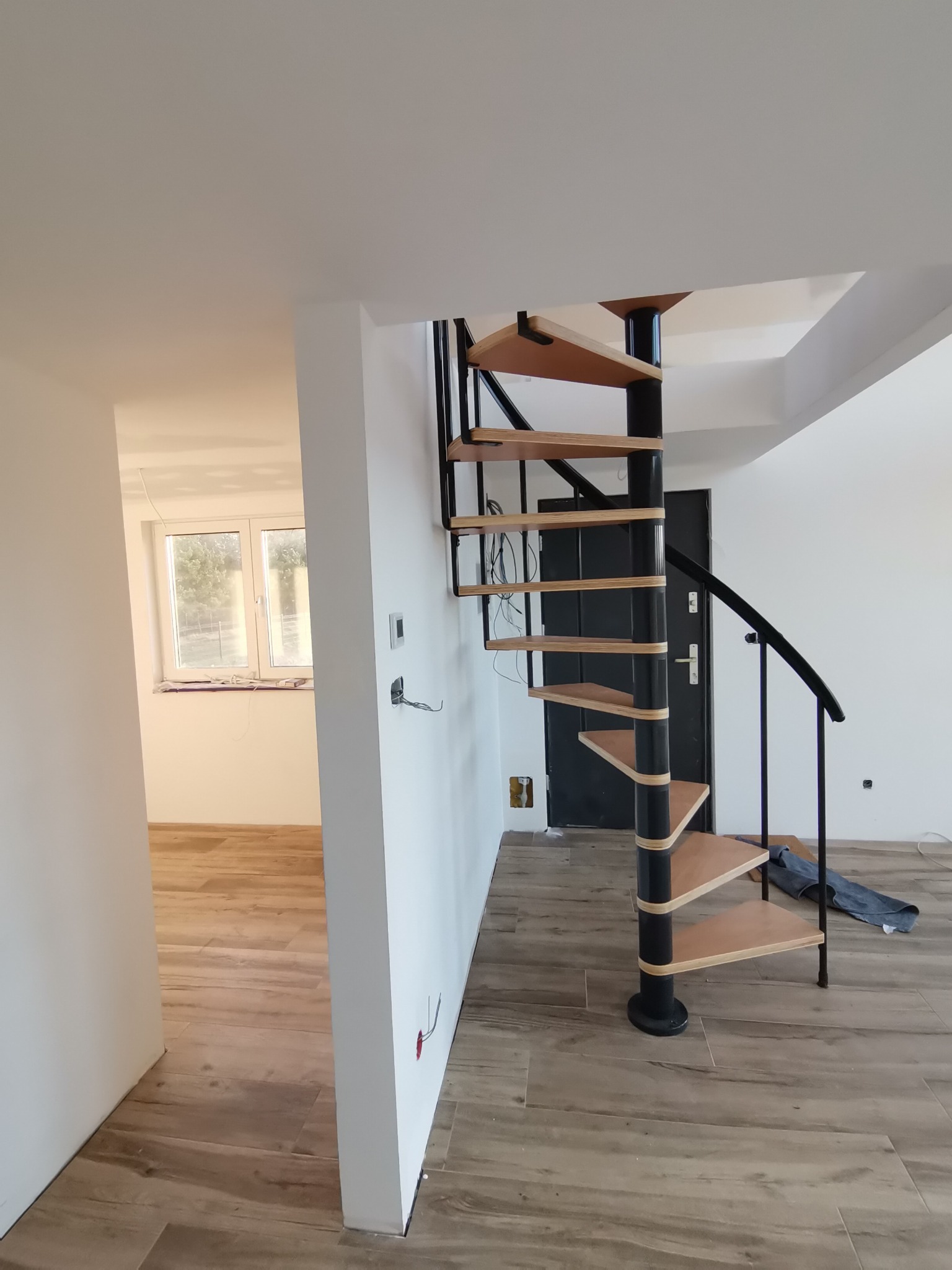Wnętrze w trakcie remontu z drewnianymi panelami podłogowymi i spiralnymi schodami z drewnianymi stopniami i czarną metalową konstrukcją, widoczne wystające kable elektryczne.