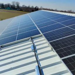 Panel fotowoltaiczny na metalowym dachu z widocznymi mocowaniami i czarnym kablem w peszlu, w tle pole i zabudowania.