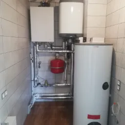 Kotłownia w jasnych płytkach, z białym piecem gazowym, zasobnikiem ciepłej wody marki Drazice, czerwonym zbiornikiem wyrównawczym i rurami w izolacji aluminiowej.