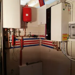 Złożona instalacja grzewcza w kotłowni z kotłem gazowym, zasobnikiem ciepłej wody i naczyniem przeponowym, z rurami w kolorach czerwonym i niebieskim, prowadzonymi wzdłuż ściany.