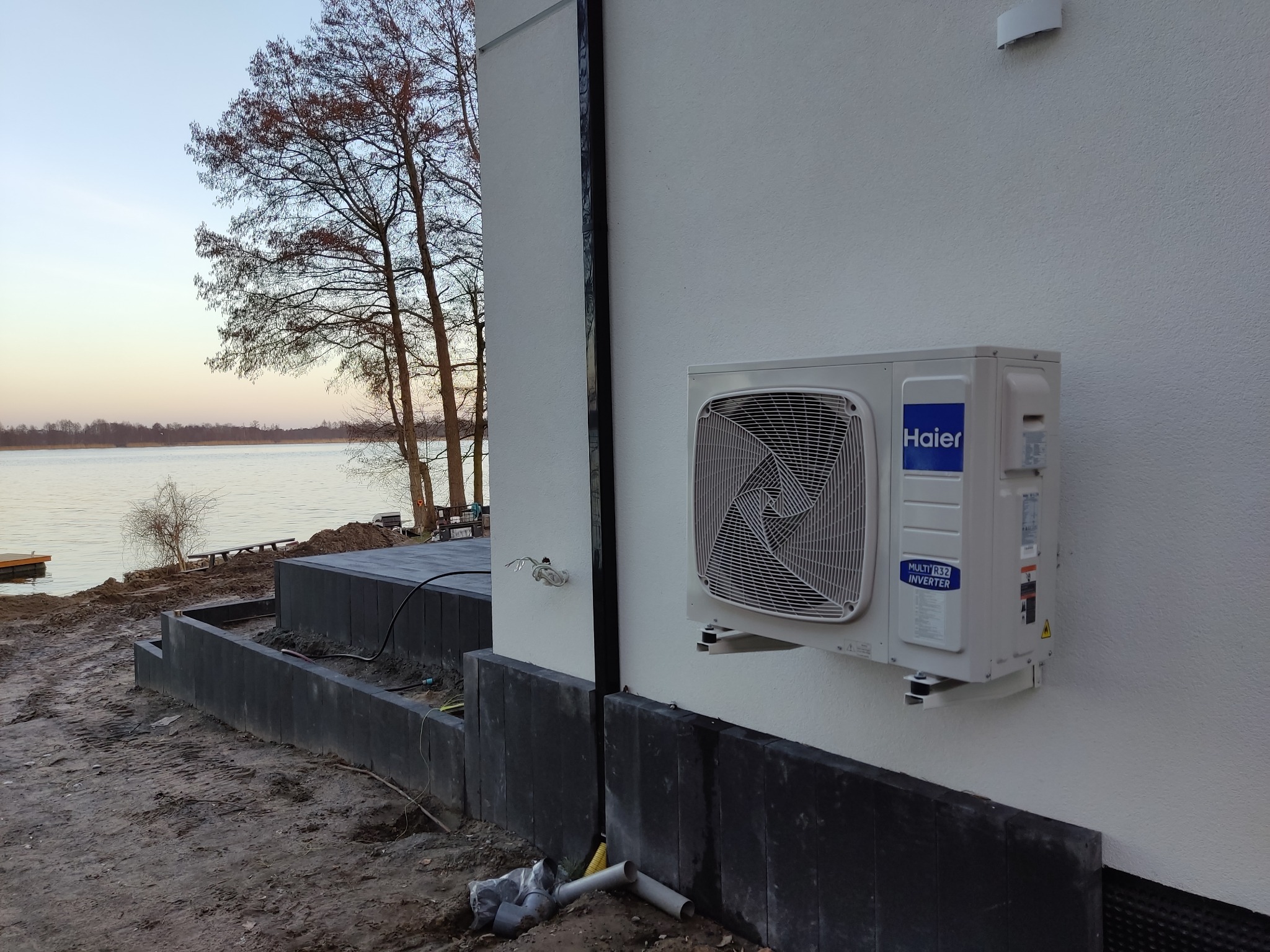 Biała jednostka zewnętrzna klimatyzacji Haier zamontowana na ścianie budynku z widokiem na jezioro o zachodzie słońca, w tle widoczne przygotowane fundamenty pod taras.