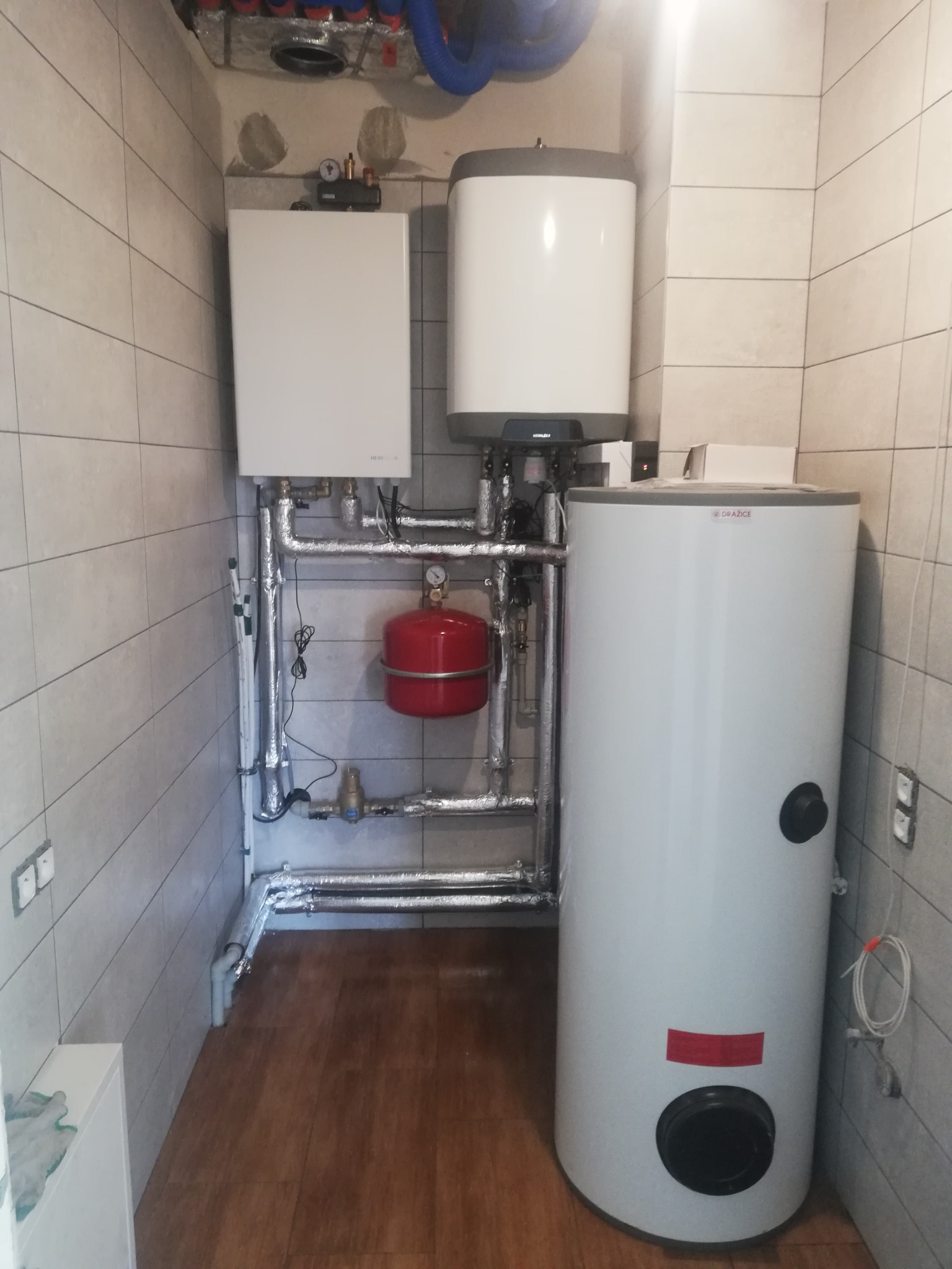 Kotłownia w jasnych płytkach, z białym piecem gazowym, zasobnikiem ciepłej wody marki Drazice, czerwonym zbiornikiem wyrównawczym i rurami w izolacji aluminiowej.