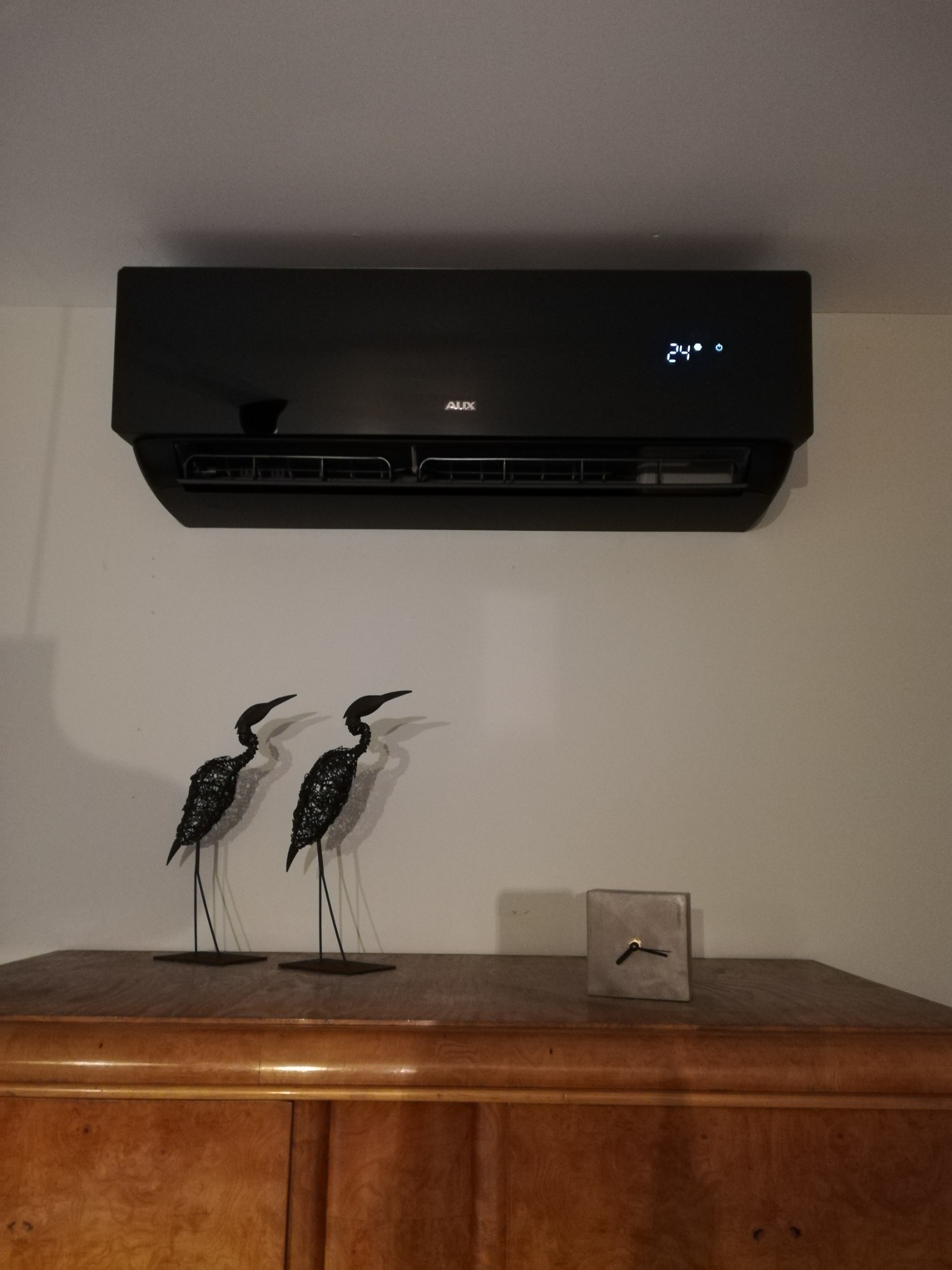 Czarny klimatyzator AUX na ścianie, wyświetlający temperaturę 24 stopnie, pod nim na komodzie dekoracyjne figurki żurawi i betonowy zegar.