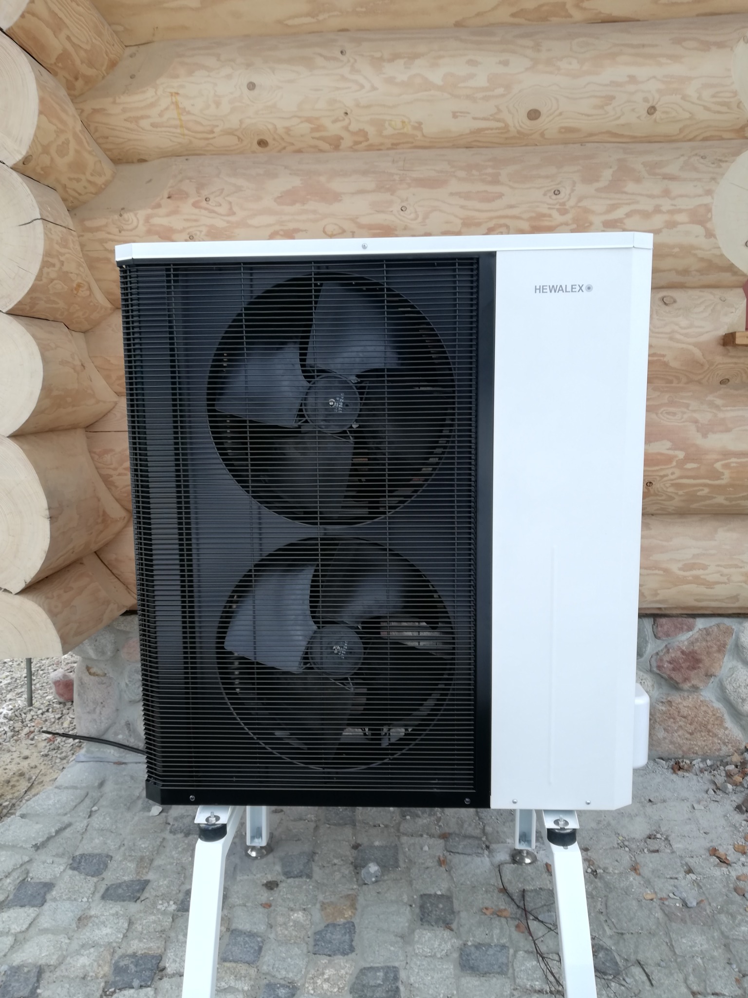 Biała pompa ciepła powietrze-woda marki Hewalex, zamontowana na zewnątrz drewnianego domu z bali, widoczne dwa wentylatory za czarną kratką.