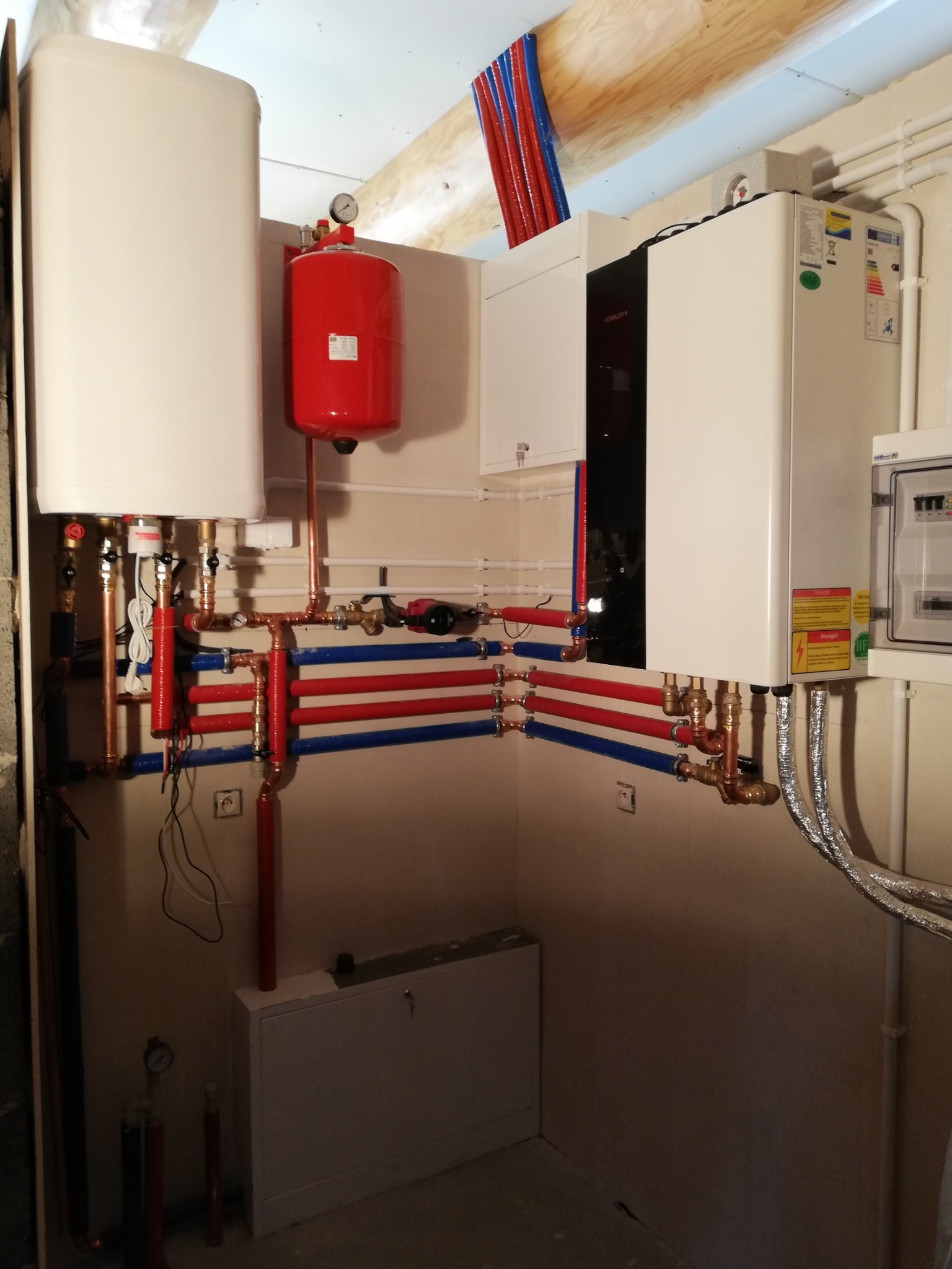 Złożona instalacja grzewcza w kotłowni z kotłem gazowym, zasobnikiem ciepłej wody i naczyniem przeponowym, z rurami w kolorach czerwonym i niebieskim, prowadzonymi wzdłuż ściany.