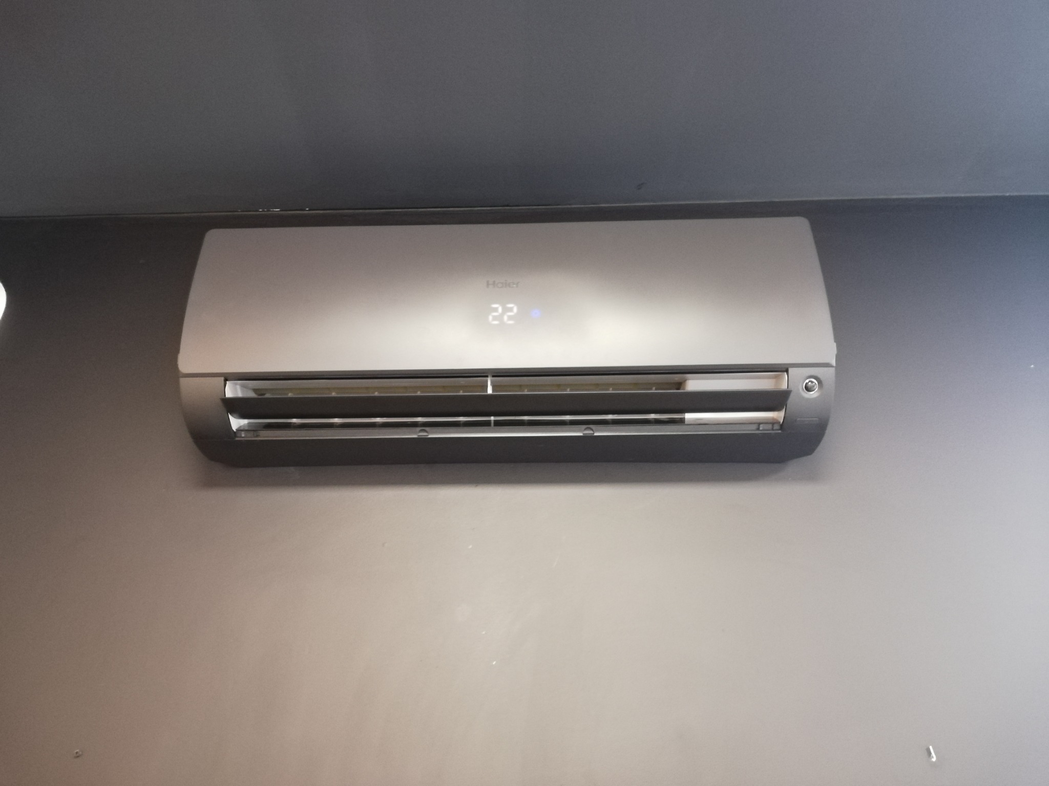 Szary klimatyzator Haier zamontowany na szarej ścianie, wyświetlający temperaturę 22 stopnie.