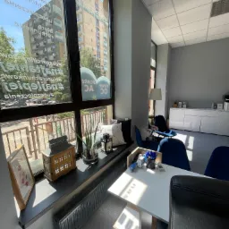 Jasne biuro z dużym oknem, widok na budynek mieszkalny z naklejkami reklamowymi na szybie, biurko z artykułami biurowymi i czarnym krzesłem, dekoracje na parapecie, niebieskie krzesła dla klientów.