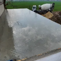 Świeżo wylany fundament pod budynek z widoczną konstrukcją szalunkową, z betoniarką w tle i zielonym polem w oddali.