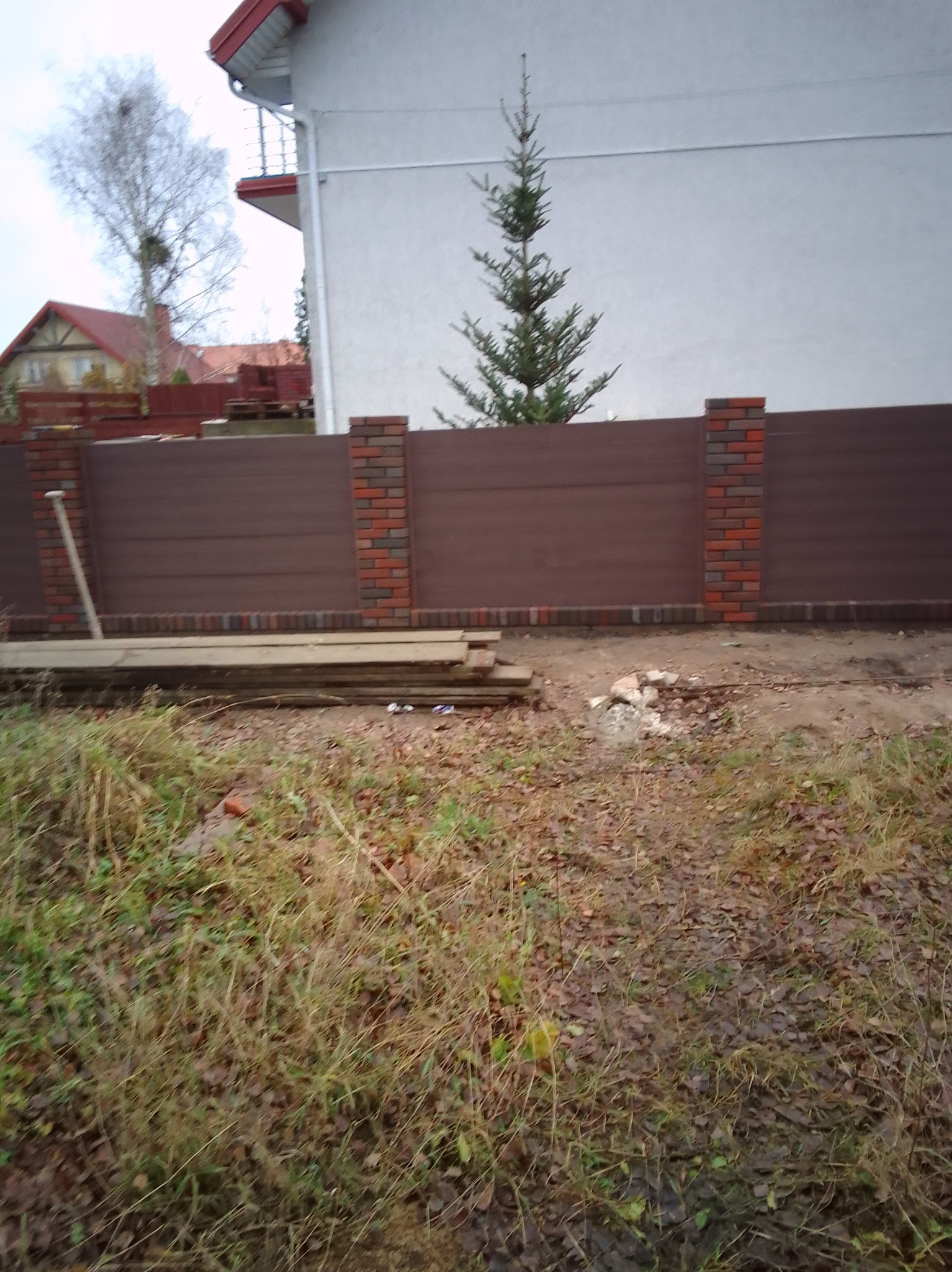 Nowoczesne ogrodzenie z brązowych paneli i ceglanych słupków, widoczne deski i narzędzie, trawa z liśćmi na pierwszym planie, dom w tle.