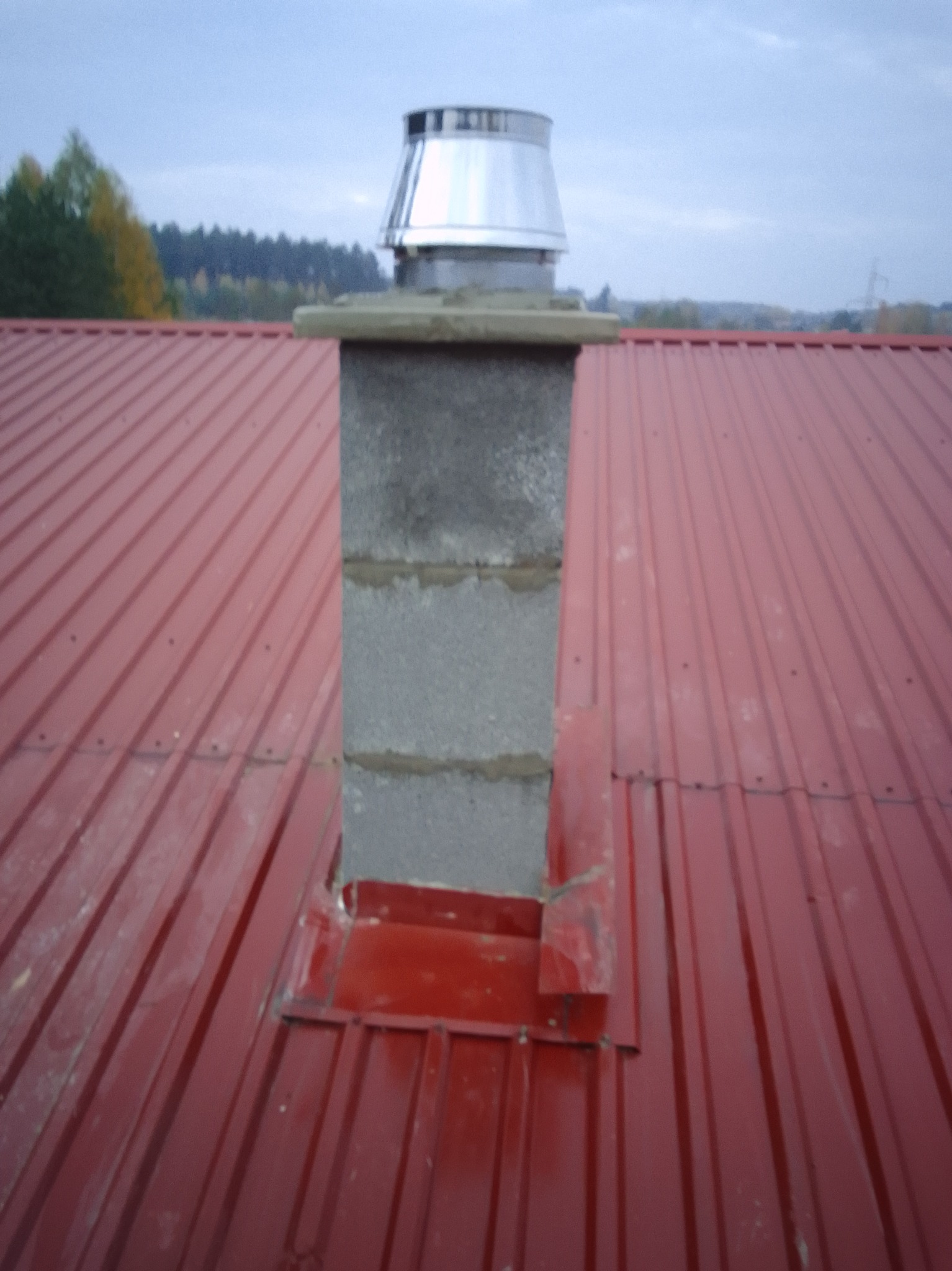 Czerwony dach z blachy trapezowej z kominem z szarego betonu i metalowym daszkiem. Widok z góry na tle drzew i pochmurnego nieba.