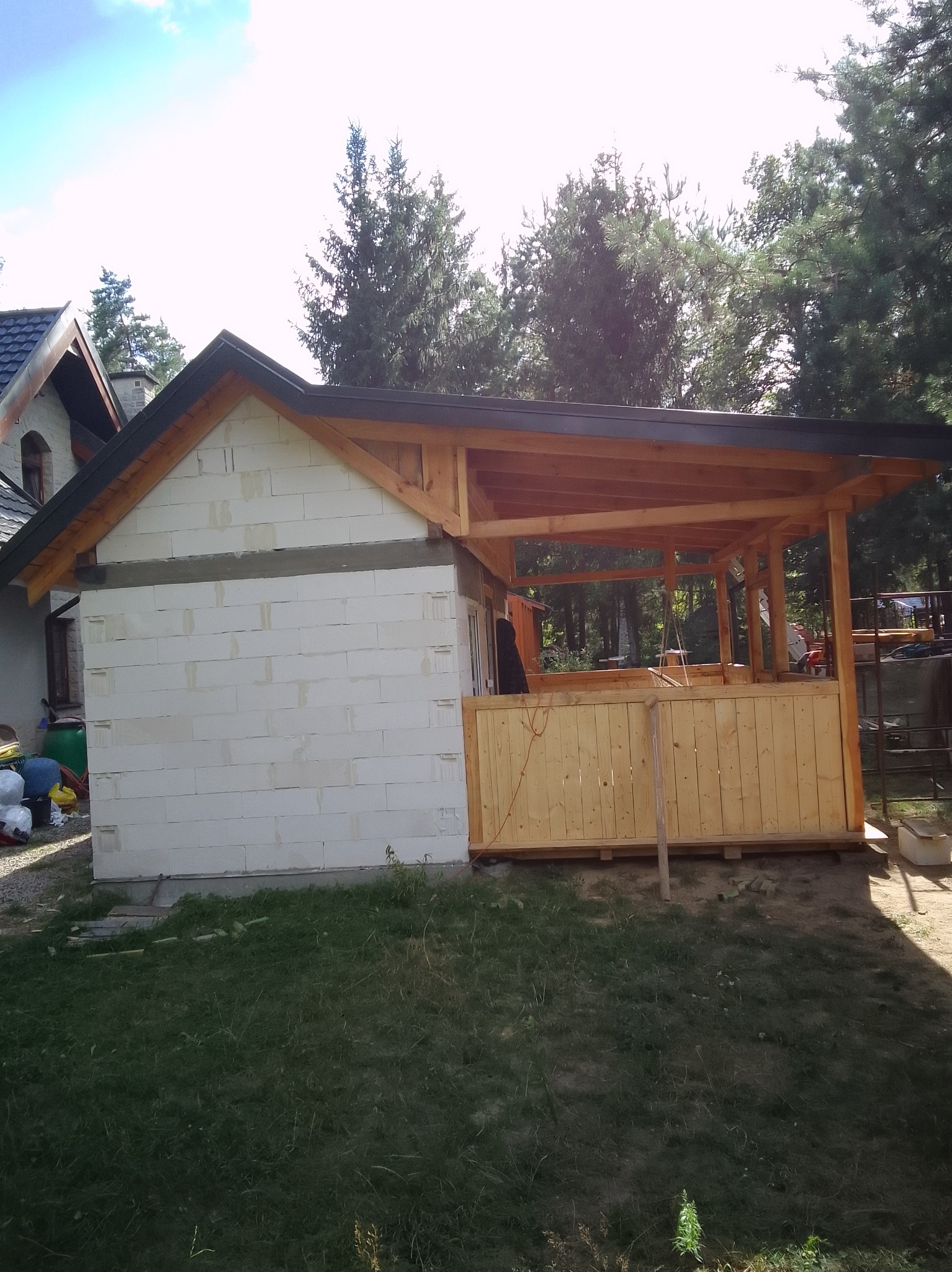 Budowa domu z białych bloczków i drewnianej werandy w otoczeniu zieleni. Widoczne elementy konstrukcyjne dachu i zadaszenia werandy. Częściowo oświetlone słońcem.