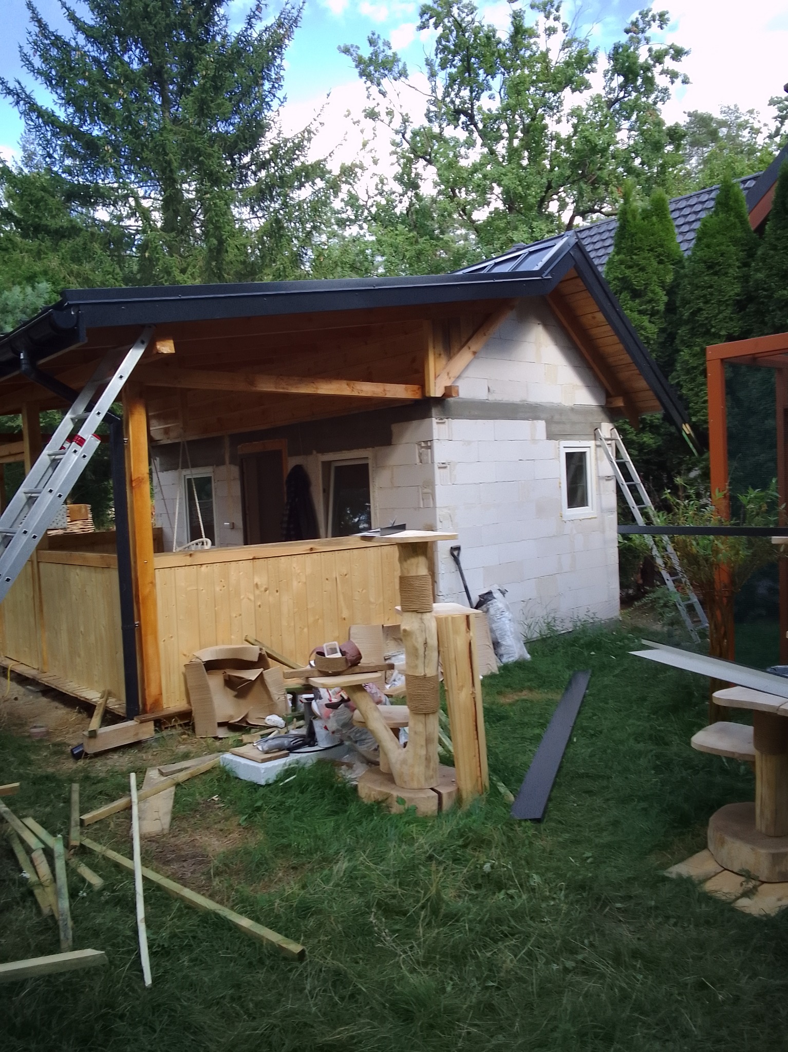 Budowa altany z drewnianą konstrukcją i dachem z blachy na tle zieleni. Widoczne materiały budowlane i drabiny. Etap prac wykończeniowych.