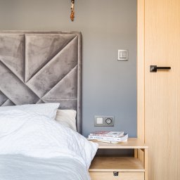 Klaudia Kwiatkowska-Wawruch - Sypialnia z szarą tapicerowaną ścianą za łóżkiem, drewniana szafka nocna z książkami i lampa wisząca z ozdobną figurką.