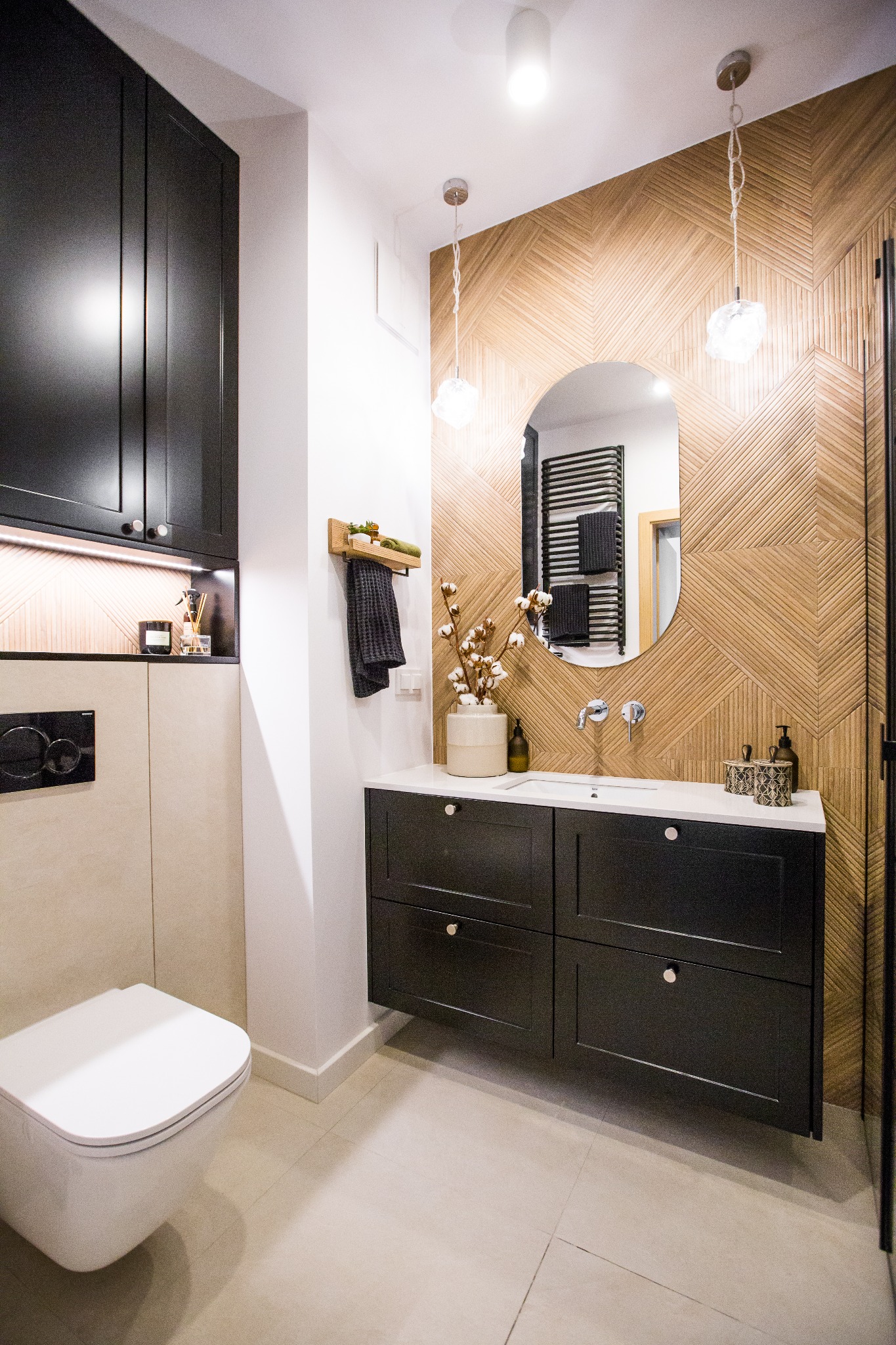Nowoczesna łazienka z drewnianą ścianą w jodełkę, czarnymi szafkami pod umywalką z białym blatem i wiszącą toaletą, oświetlona lampami wiszącymi z efektem kryształu.