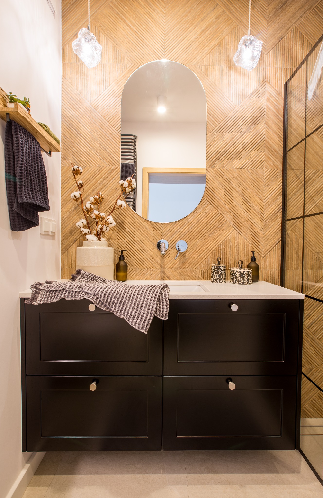 Nowoczesna łazienka z drewnianą mozaiką na ścianie, owalnym lustrem, czarną szafką pod umywalką z marmurowym blatem, bawełnianymi gałązkami w wazonie i szklaną kabiną prysznicową.