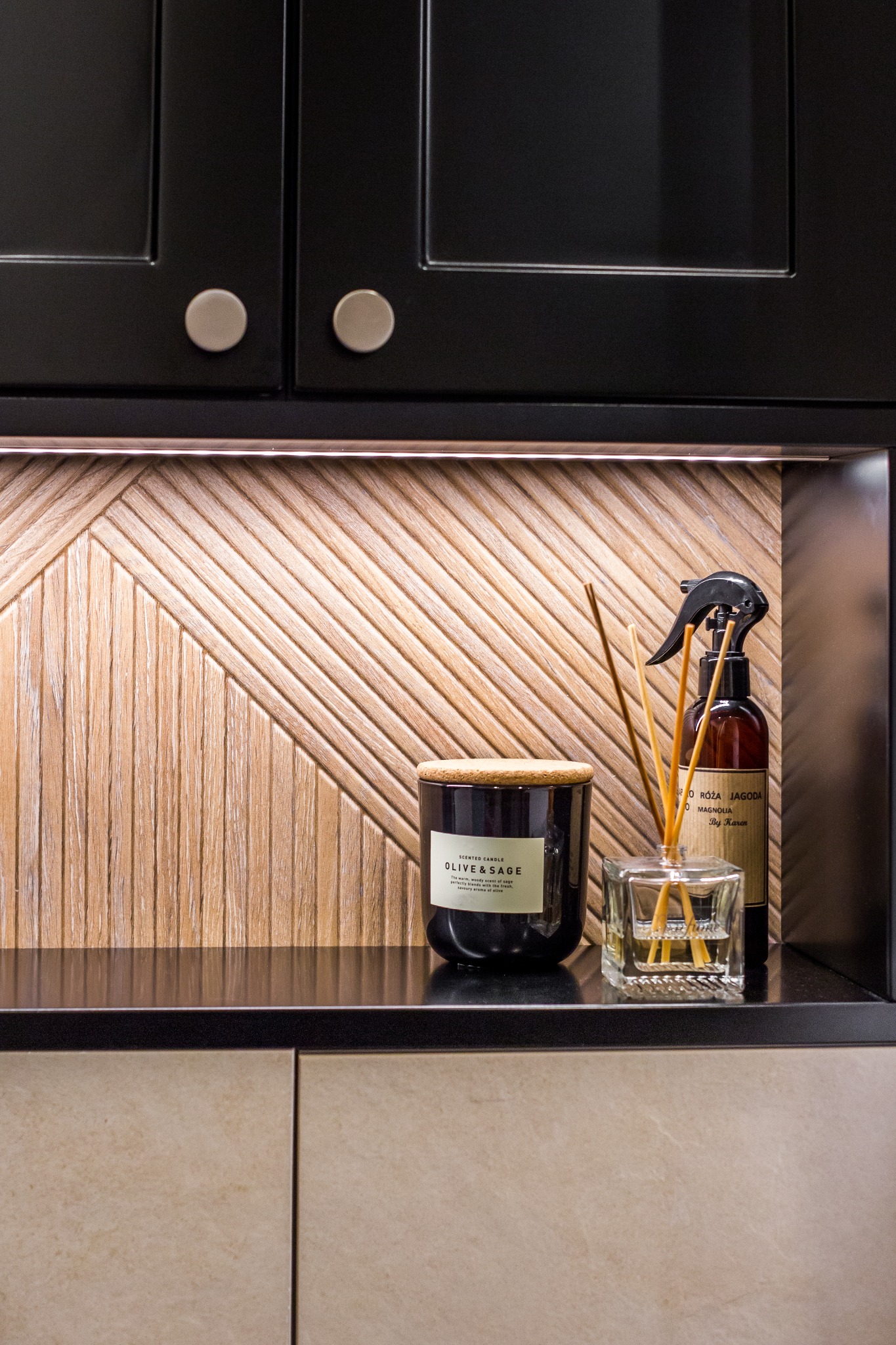 Dekoracyjny panel ścienny z drewna w łazience, podświetlony LED, z czarnymi szafkami i akcesoriami zapachowymi: świecą Olive & Sage, dyfuzorem zapachowym i sprayem By Karen.