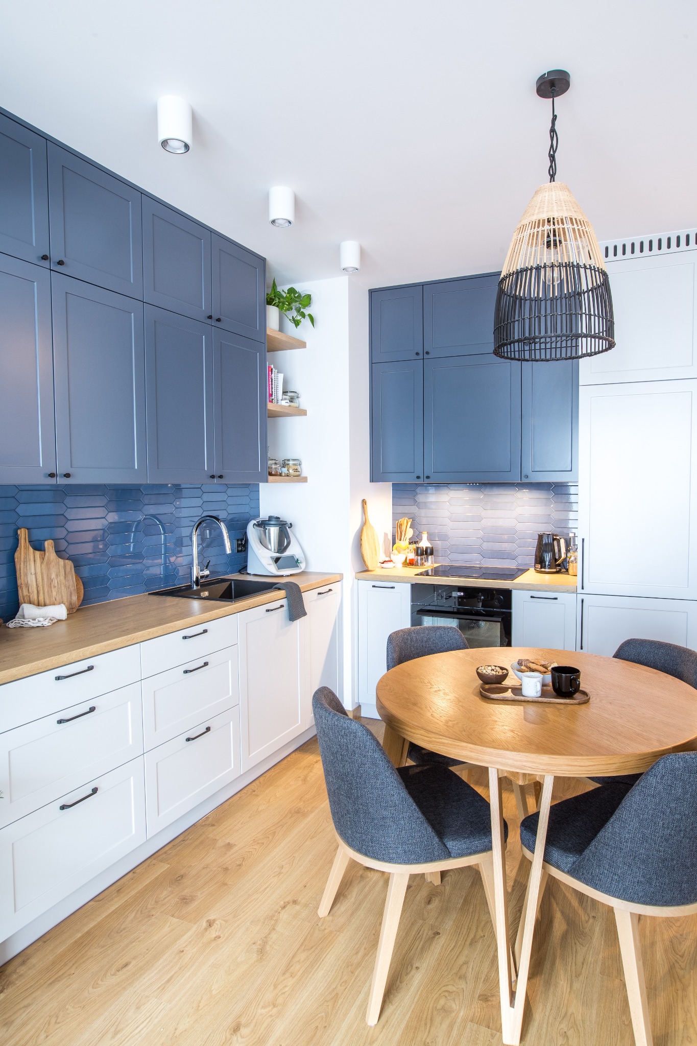 Nowoczesna kuchnia z niebieskimi szafkami górnymi, białymi dolnymi, drewnianym blatem i okrągłym stołem z szarymi krzesłami, oświetlona lampą z wikliny.