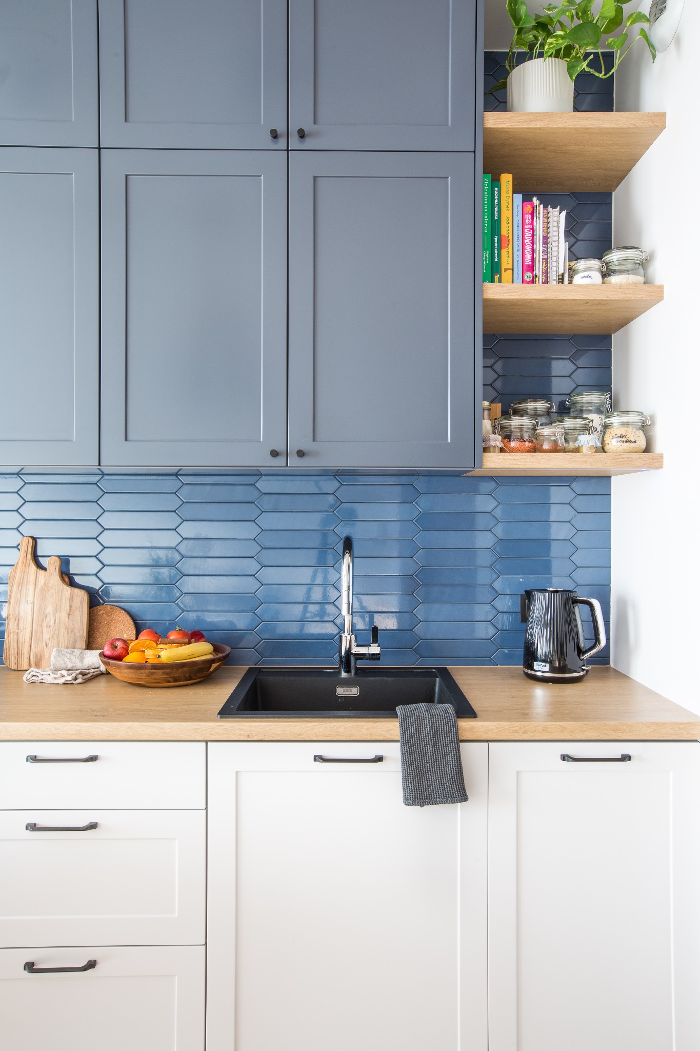 Nowoczesna kuchnia z szarymi szafkami górnymi, białymi dolnymi, drewnianym blatem i niebieskimi płytkami ułożonymi w jodełkę. Na blacie czarny zlew, bateria, czajnik i misa z owocami. Półki...